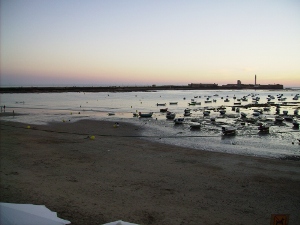 Playa la Caleta con el sol poniéndose. En primer plano se ven barquitas y al fondo el castillo